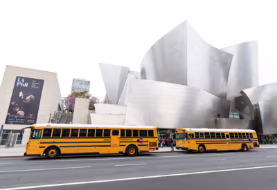White concert hall with curved stone façade and two yellow buses parked in front, billboard on left