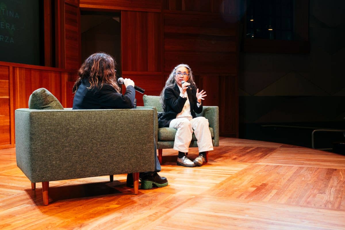 Two women seated on stage during an interview, one speaking into a microphone.