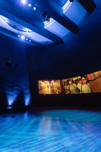 Dark theater studio with blue lighting, empty stage, and a wall screen showing people and colorful panels