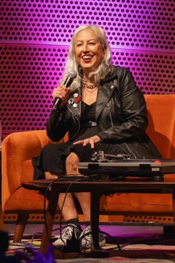 Smiling older woman in a black leather jacket speaks into a microphone on an orange stage with a panel table.