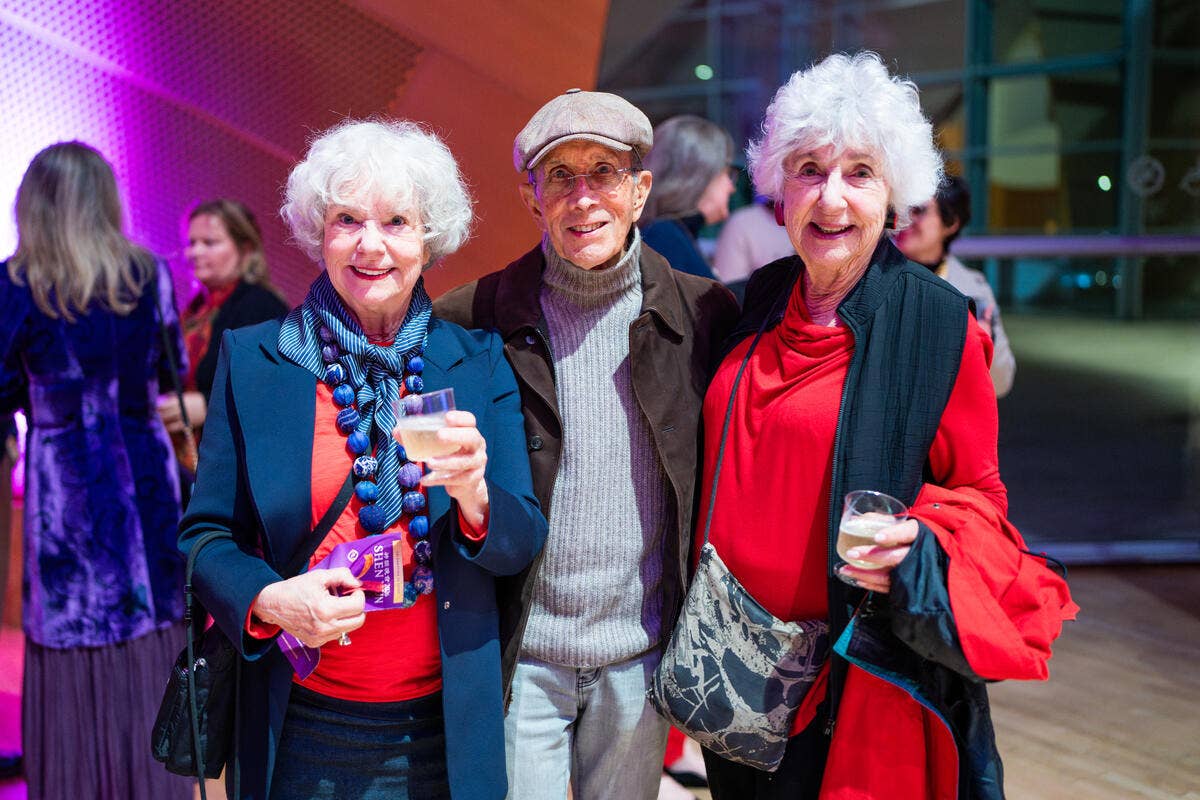 Three older adults smiling at an event, holding drinks and books in a brightly lit indoor hall.