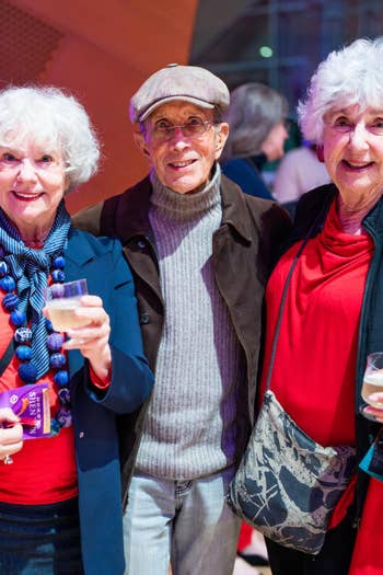 Three older adults smiling at an event, holding drinks and books in a brightly lit indoor hall.