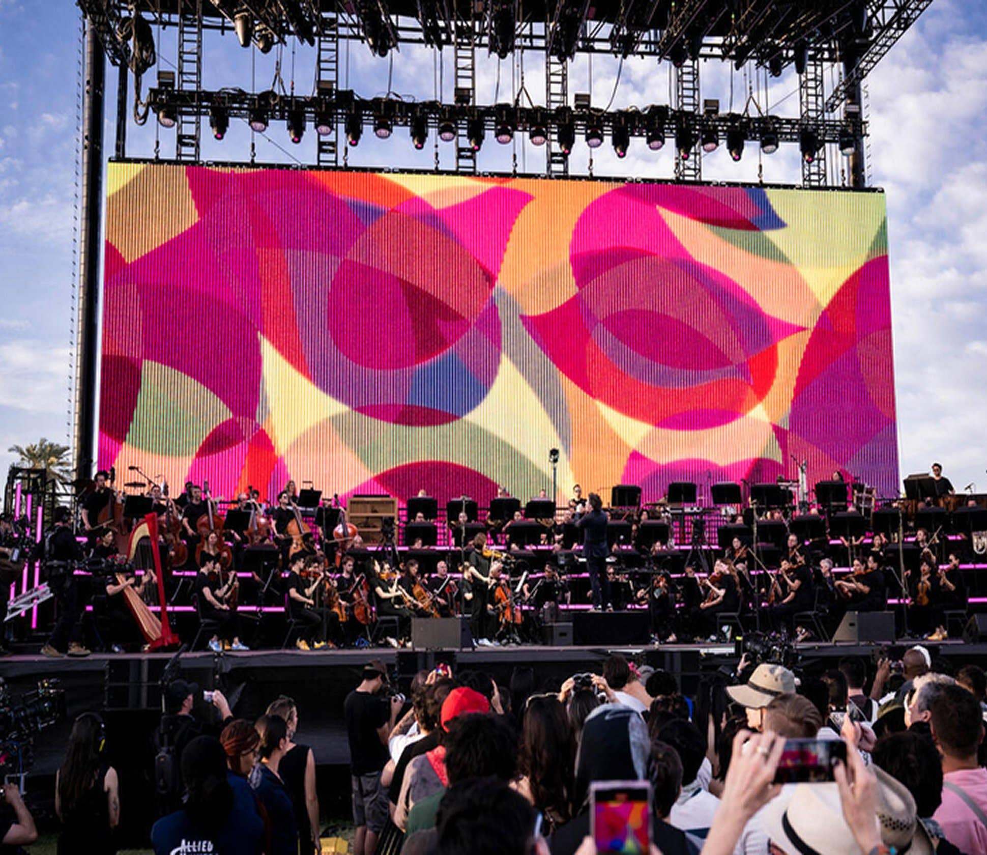 Orchestra on an outdoor stage beneath scaffolding lights, performing before a colorful abstract LED screen.