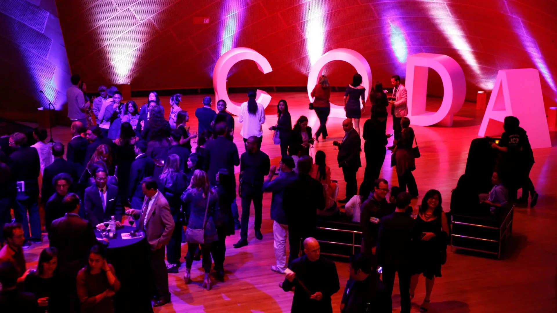 Conference attendees walk on stage under colorful red and purple lighting, with large illuminated letters “G D A” behind them.