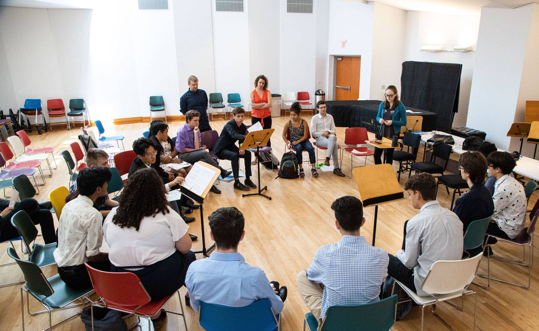 People seated in a circle during a workshop, some reading sheet music, with speakers and chairs in a bright room