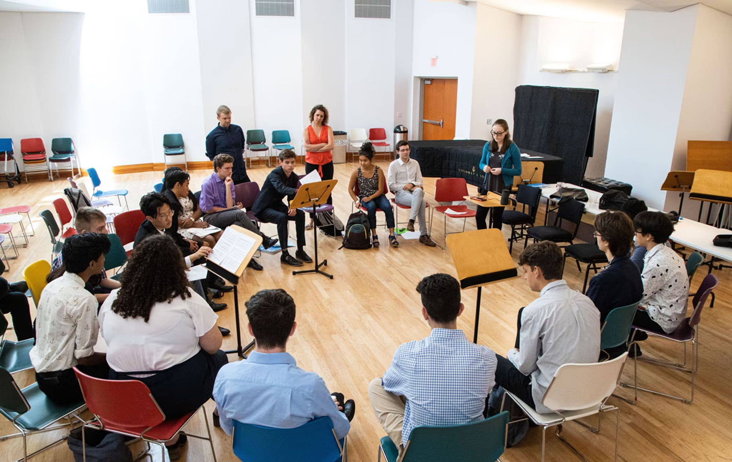 People seated in a workshop circle, listening as a facilitator speaks with papers and laptops on stands.