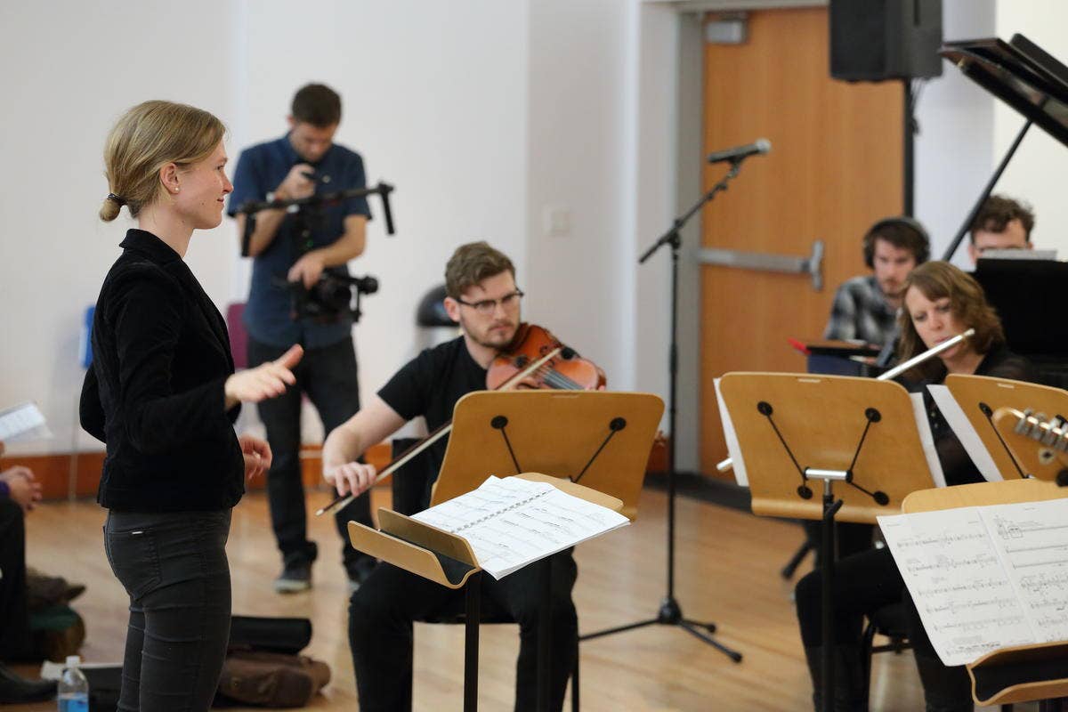 Woman conducts while musicians play violin and flute in a rehearsal room with sheet music stands and microphones.