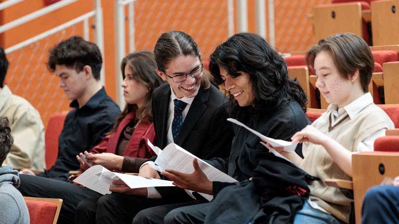 Students in a lecture hall smiling and reading printed papers in red-cushioned seats