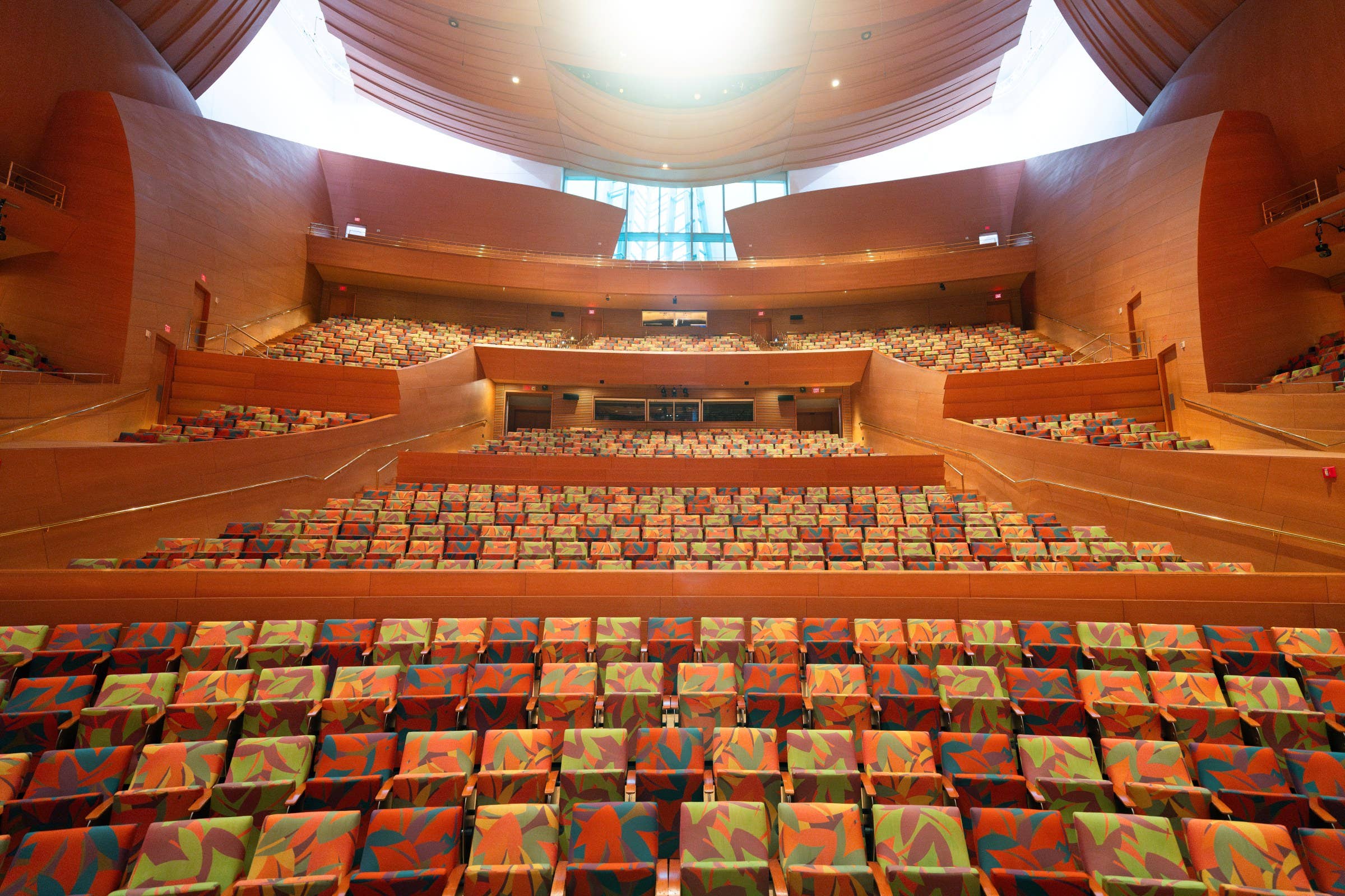 Empty auditorium with colorful patterned seats, tiered balconies, and a curved wood ceiling with skylight.