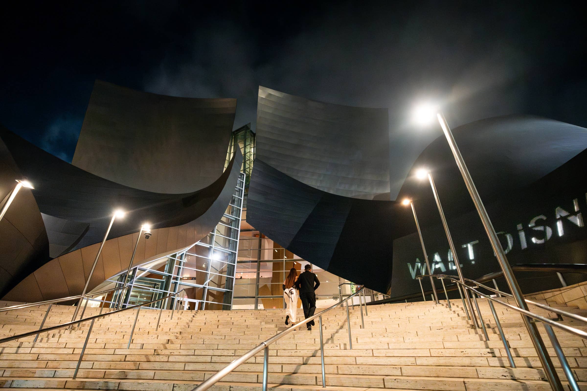Night view of modern museum steps, curved dark facades, and bright spotlights with two people walking.