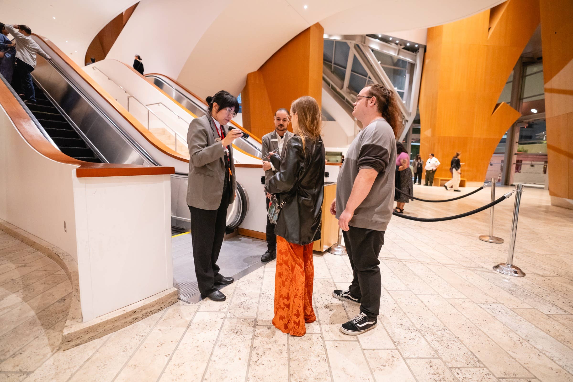 Conference attendees inspect museum-style exhibit near escalators and an orange wood staircase wall.
