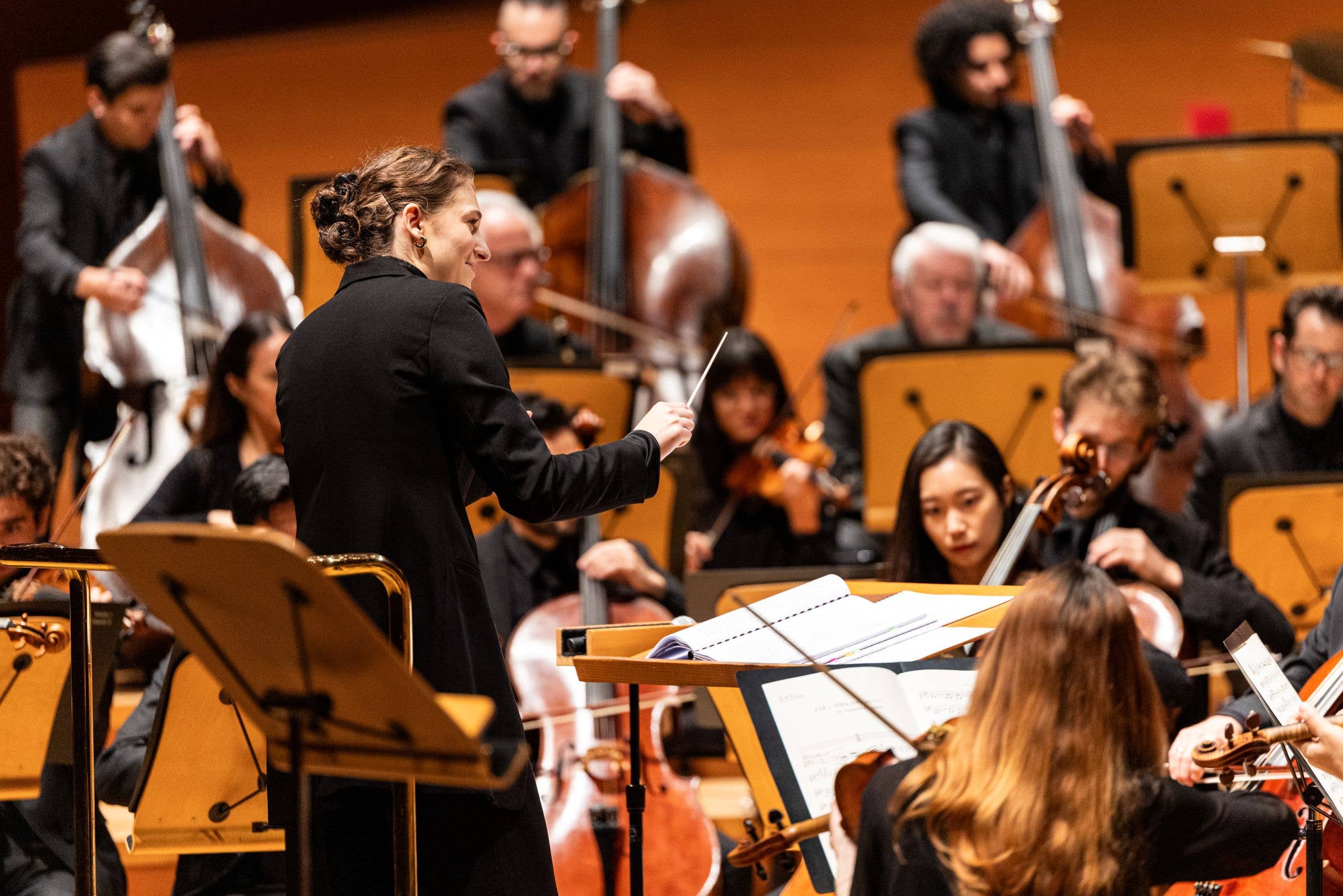 Orchestra rehearsal with musicians playing violins and cellos on a concert stage, conductor in front.