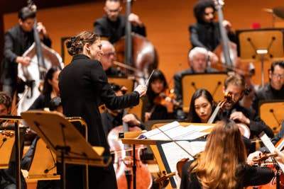 Orchestra rehearsal with musicians playing violins and cellos on a concert stage, conductor in front.