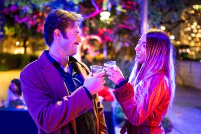 Man and woman smile at an outdoor night event, clinking glasses with colorful purple and red lighting.