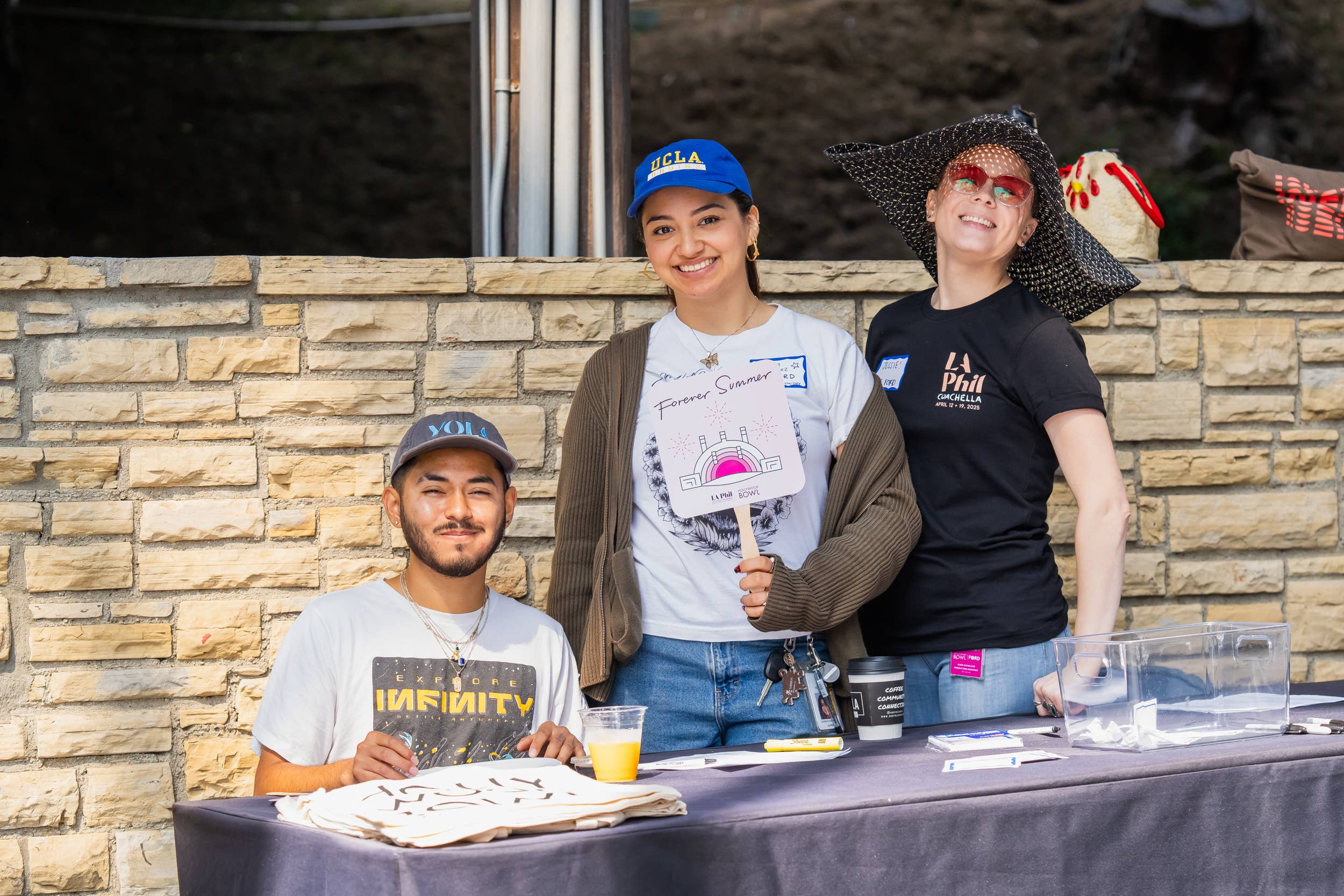 Three people smiling at an outdoor event table; one holds a “Forever Summer” fan, another wears a UCLA cap.