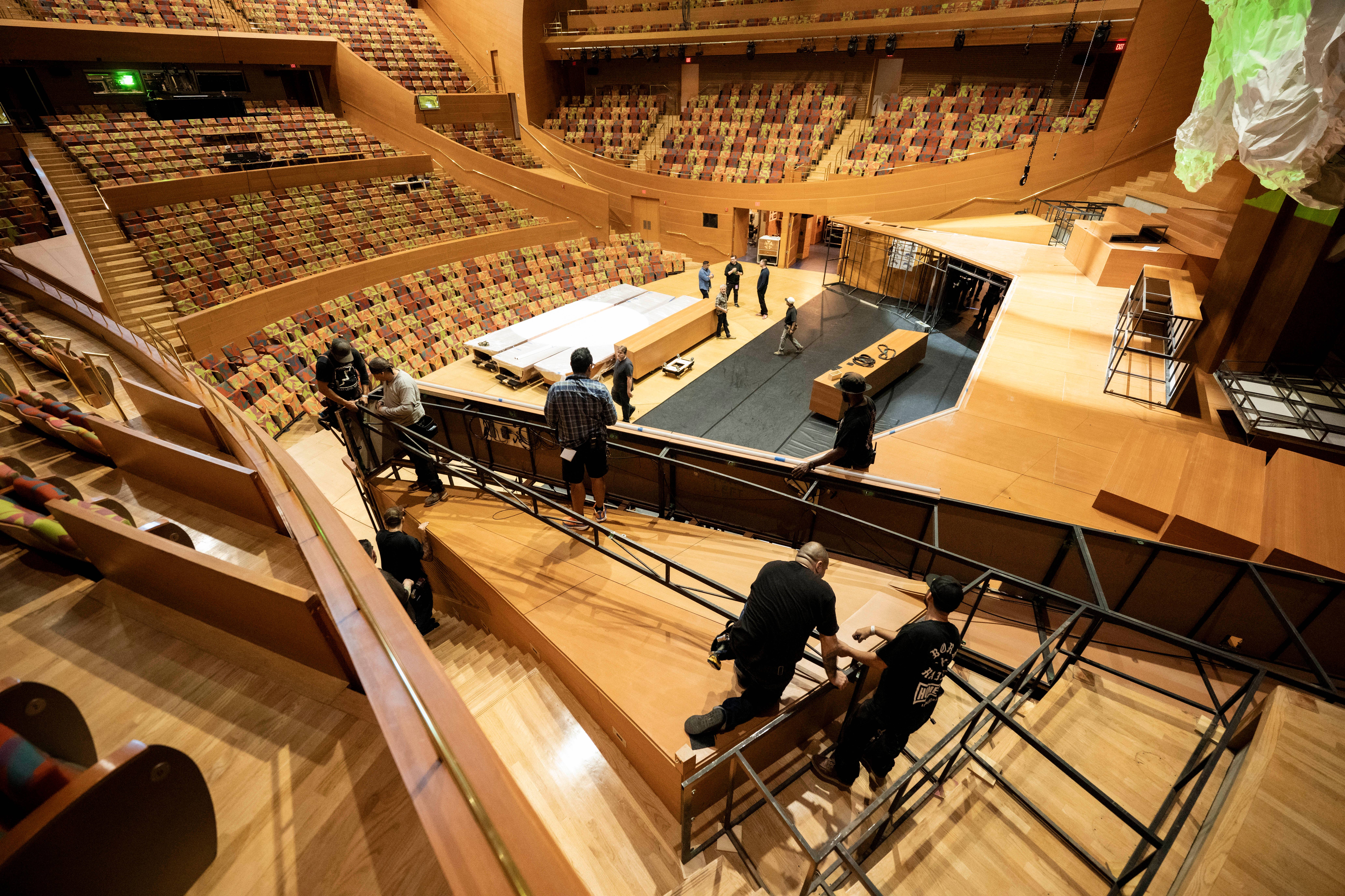 Audience seating filled with colorful chairs surrounds a stage where people walk on a gray floor.