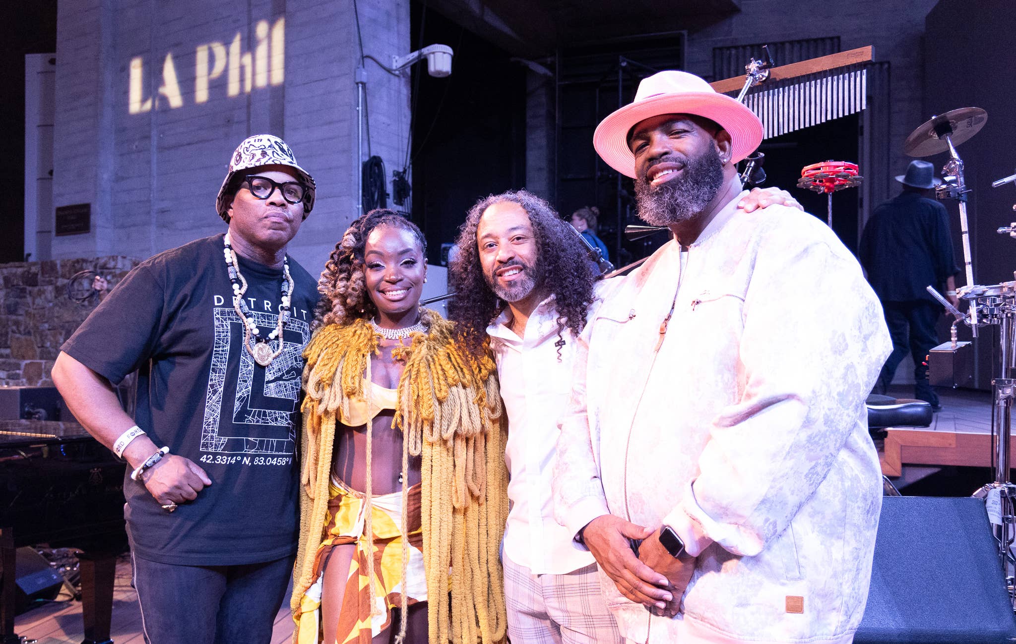 Four performers pose at a music venue stage, including a smiling man in a pink hat and white outfit.