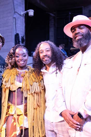 Four performers pose at a music venue stage, including a smiling man in a pink hat and white outfit.