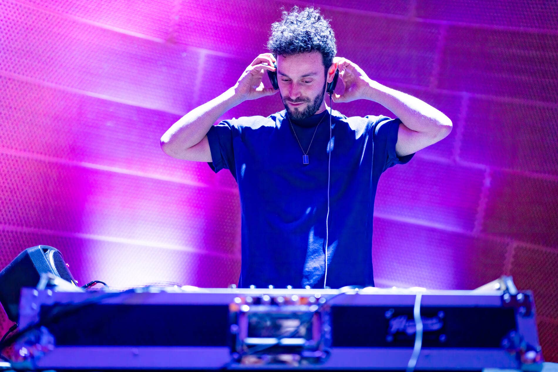 DJ with curly hair adjusts headphones while performing behind a mixing console under purple stage lights.