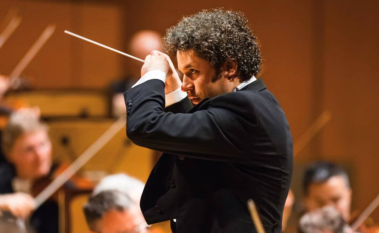 Curly-haired conductor in dark suit holding a baton, leading an orchestra rehearsal onstage