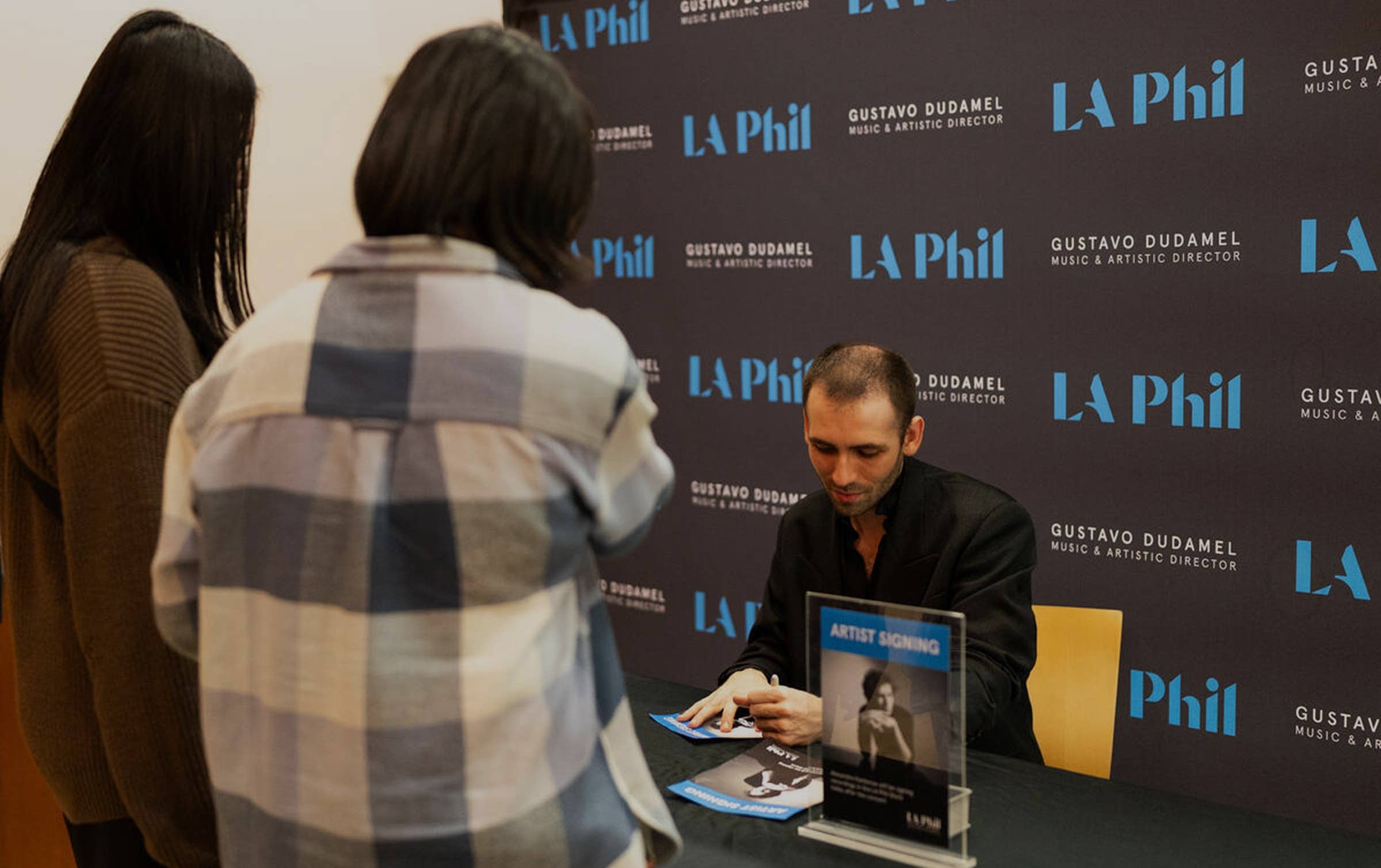 Man in a dark jacket signs an album while a woman stands in front at a LA Phil backdrop event.