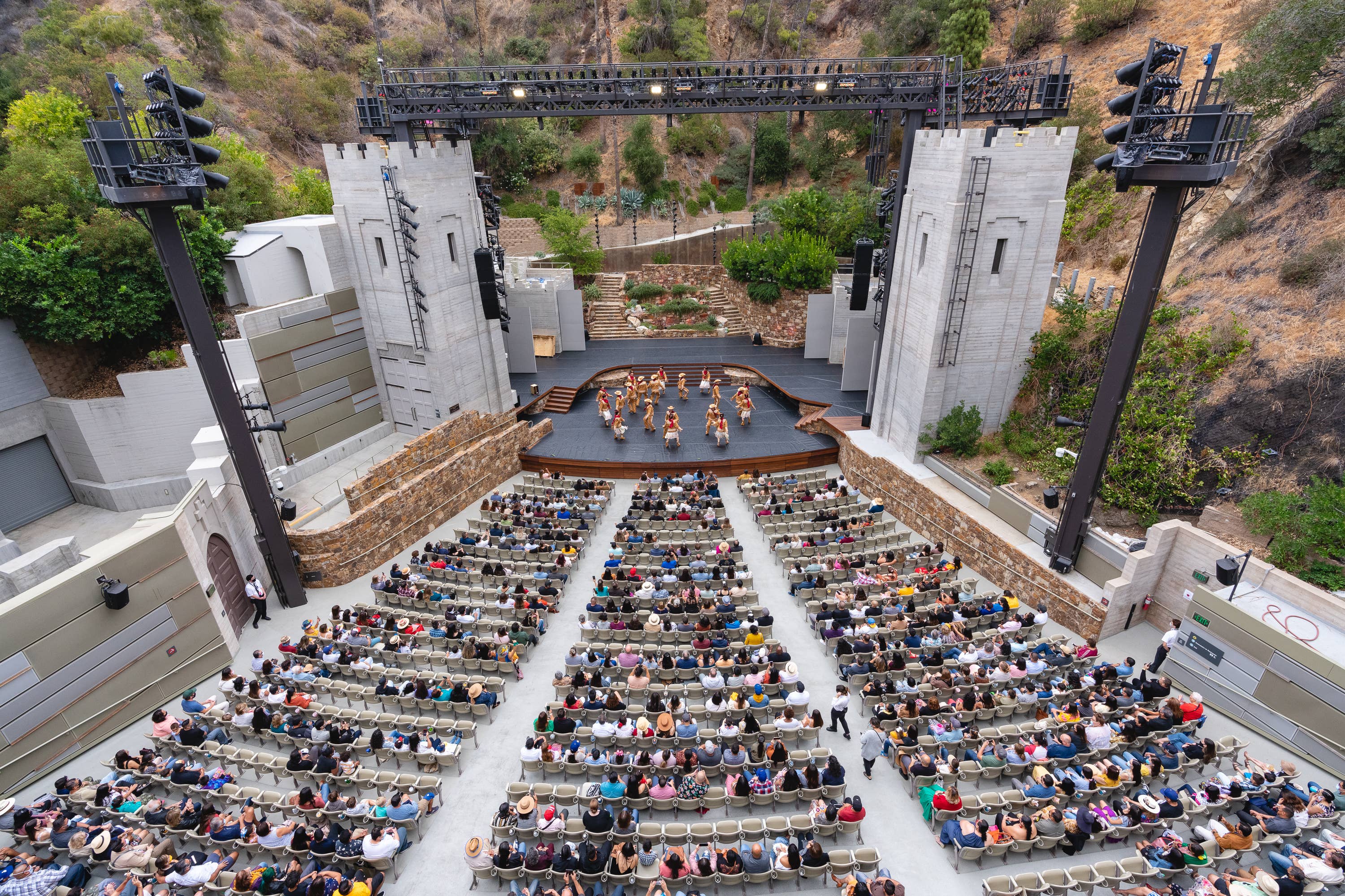 Crowd seated in outdoor amphitheater as performers dance on stage framed by truss lighting and white walls