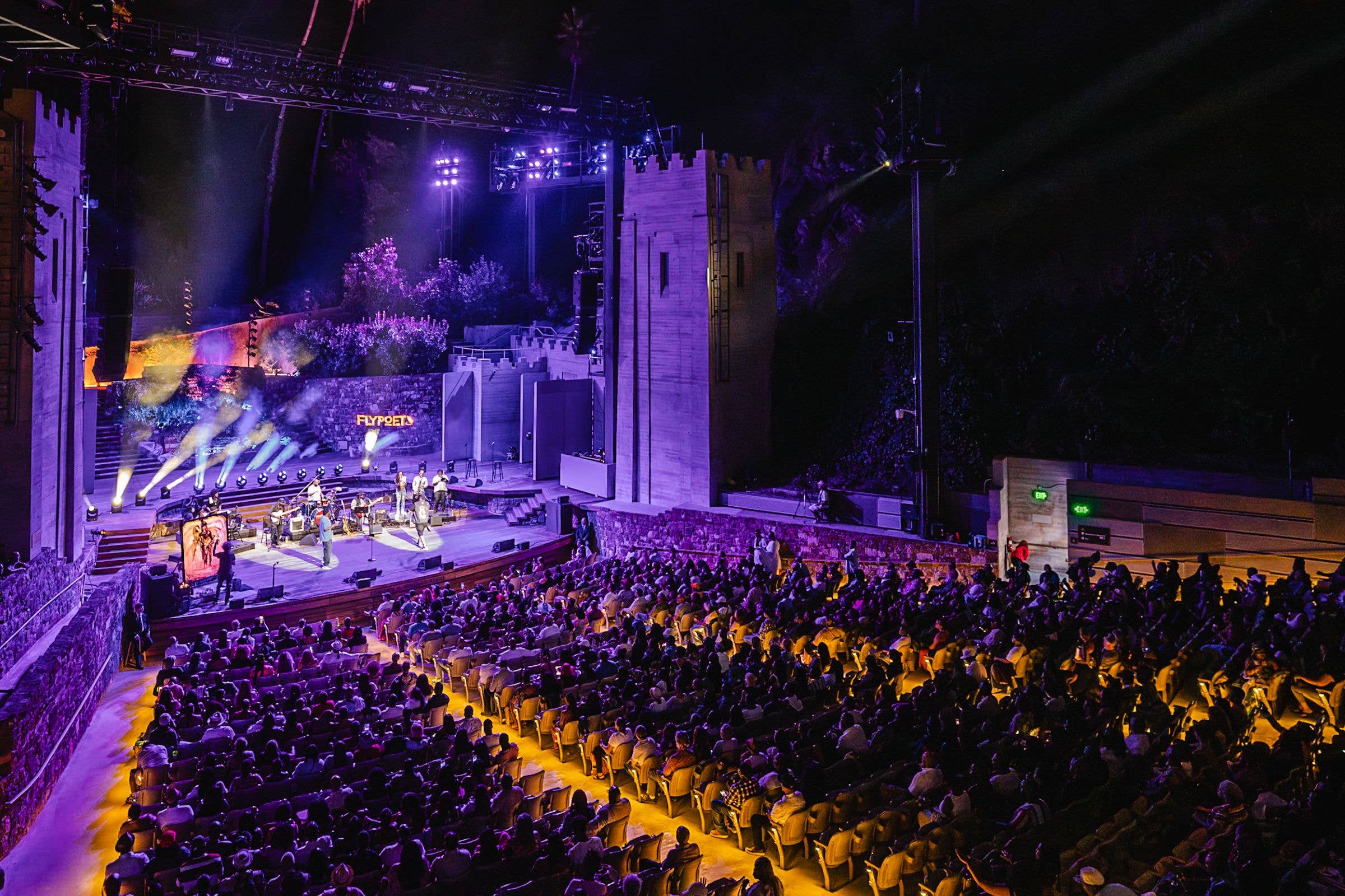 Concert hall stage with purple lighting, smoke effects, fireworks, and a crowd seated in rows.