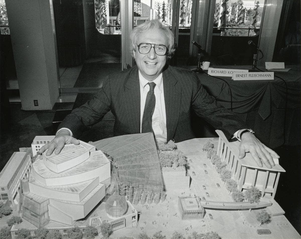 Smiling man in suit and glasses at a conference table behind a detailed architectural scale model