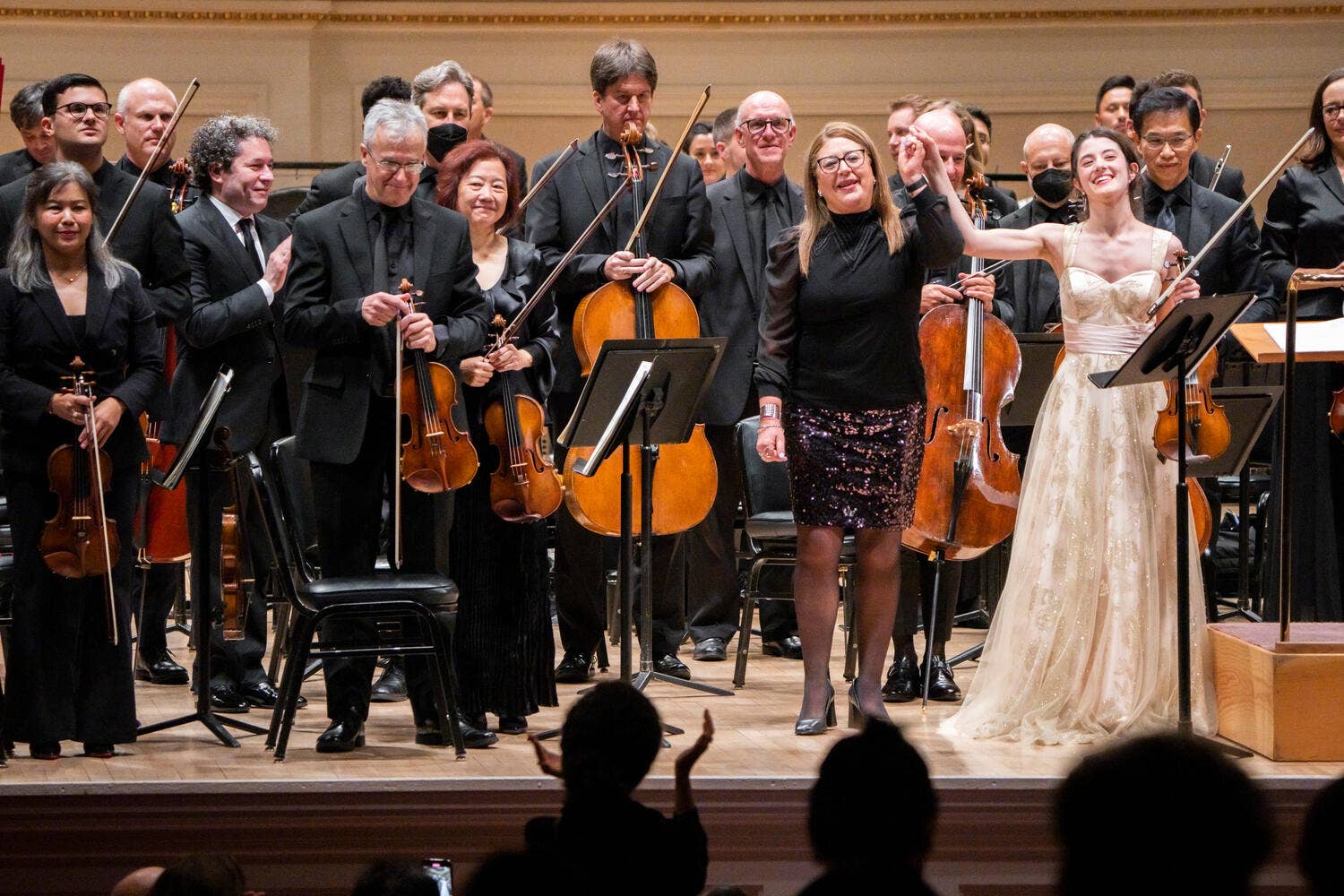 Orchestra members perform on stage with violins and cello, smiling conductor at center, audience silhouettes in foreground