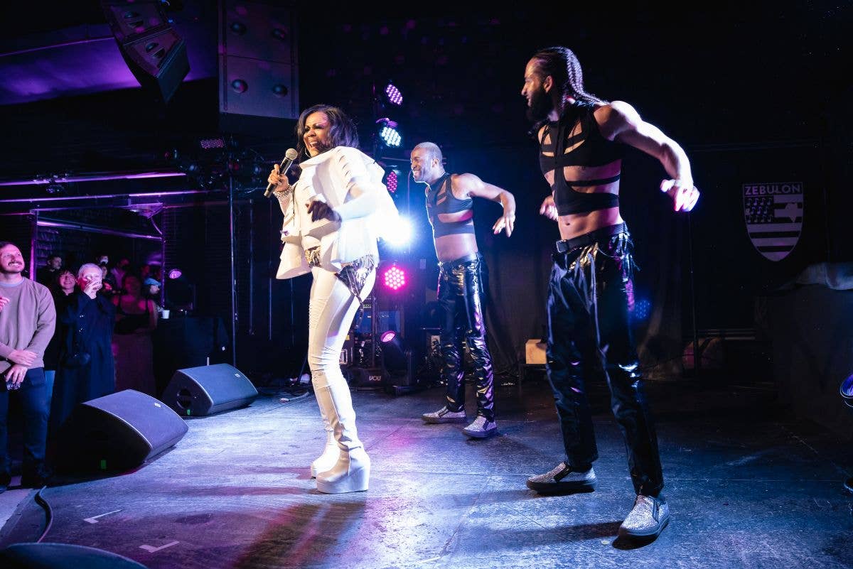 Singer in white suit performs with dancers on stage under purple LED lights and spotlights, holding a microphone.