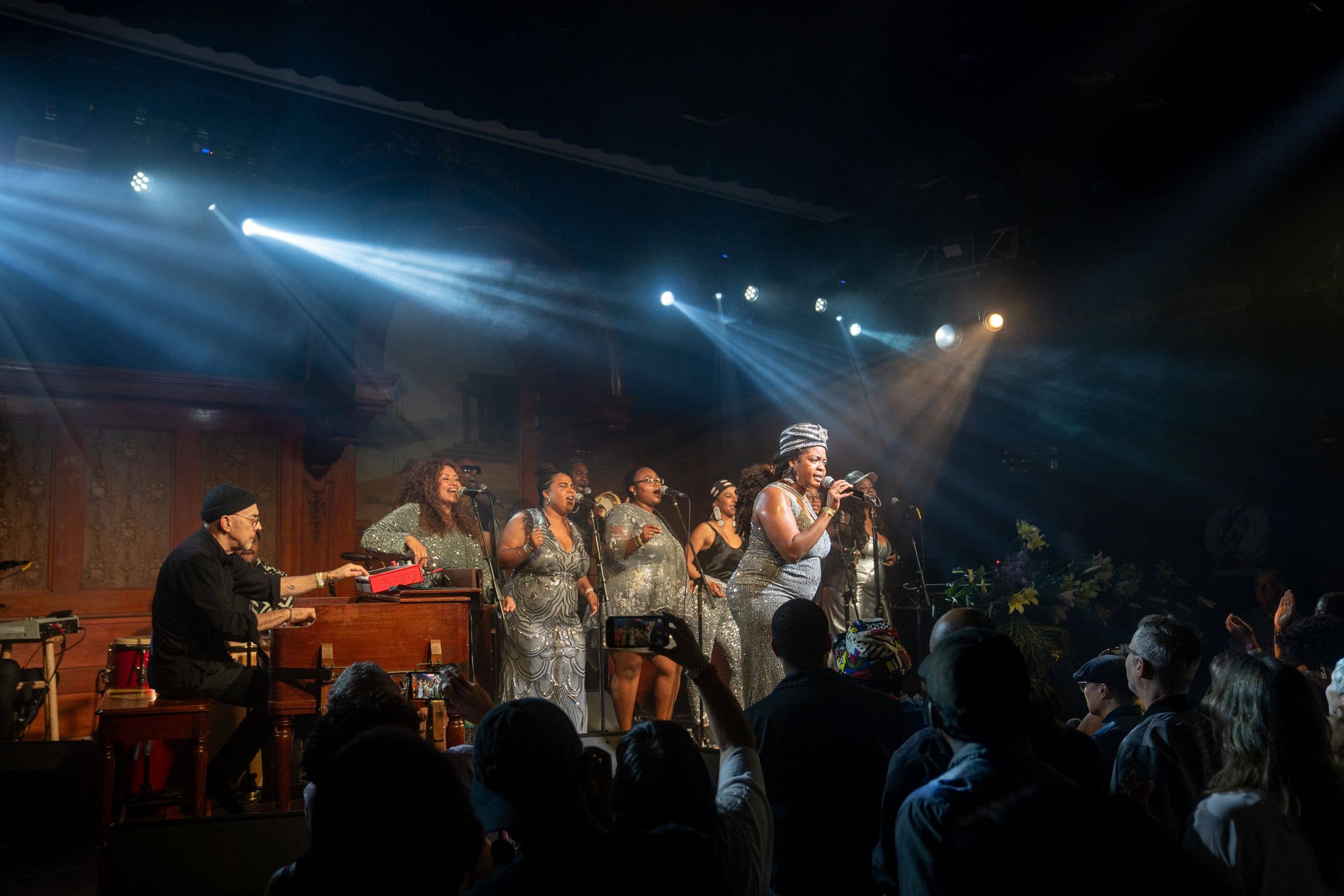 Choir singers in silver dresses perform onstage under blue spotlights, with an audience facing microphones and music stand