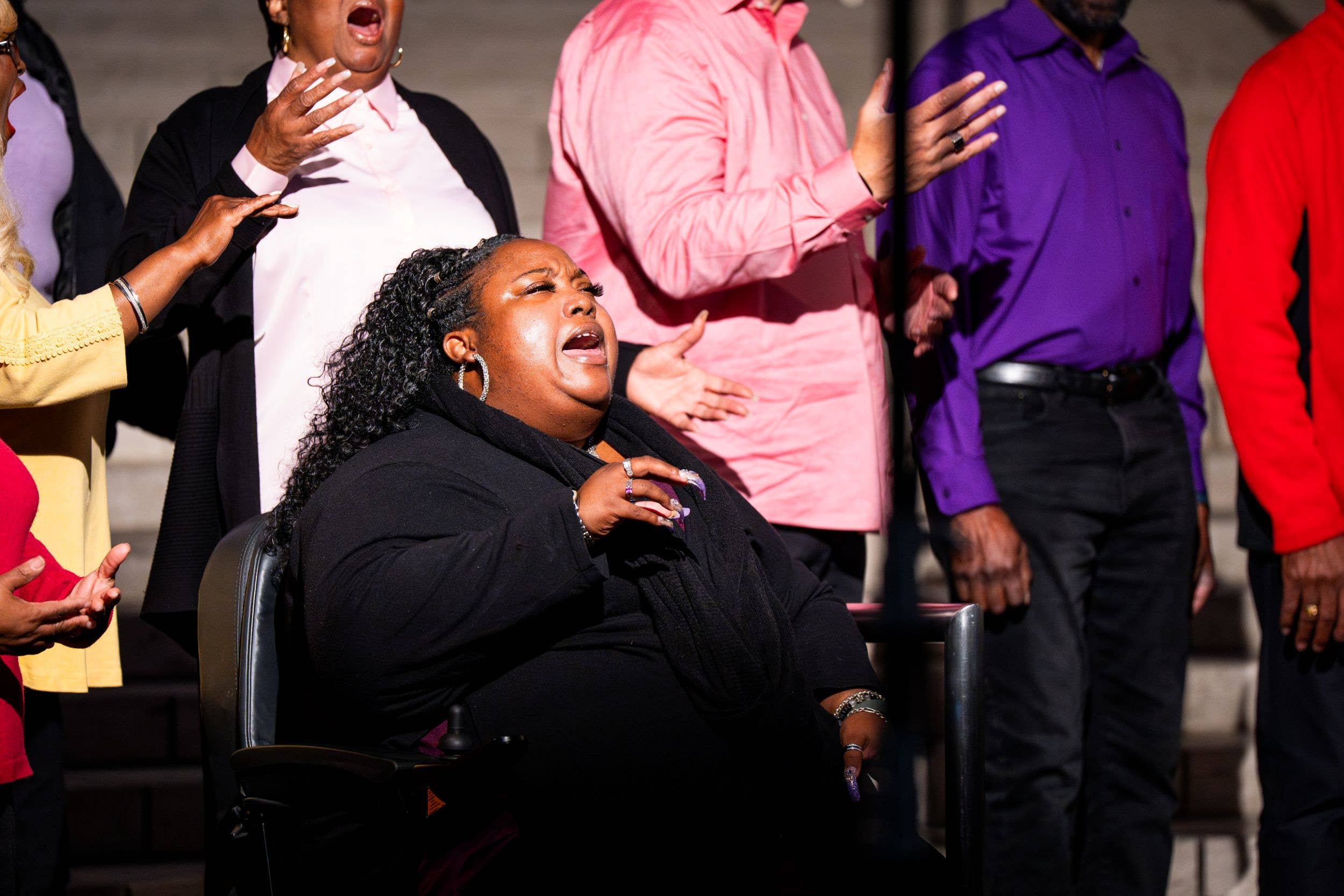 Woman seated and speaking with expressive gestures while a man and crowd stand behind her under bright lights