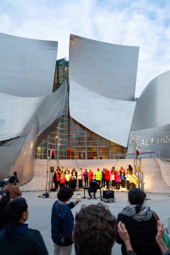 Crowd gathers in front of modern curved museum building with large white stone facade and steps outdoors at dusk