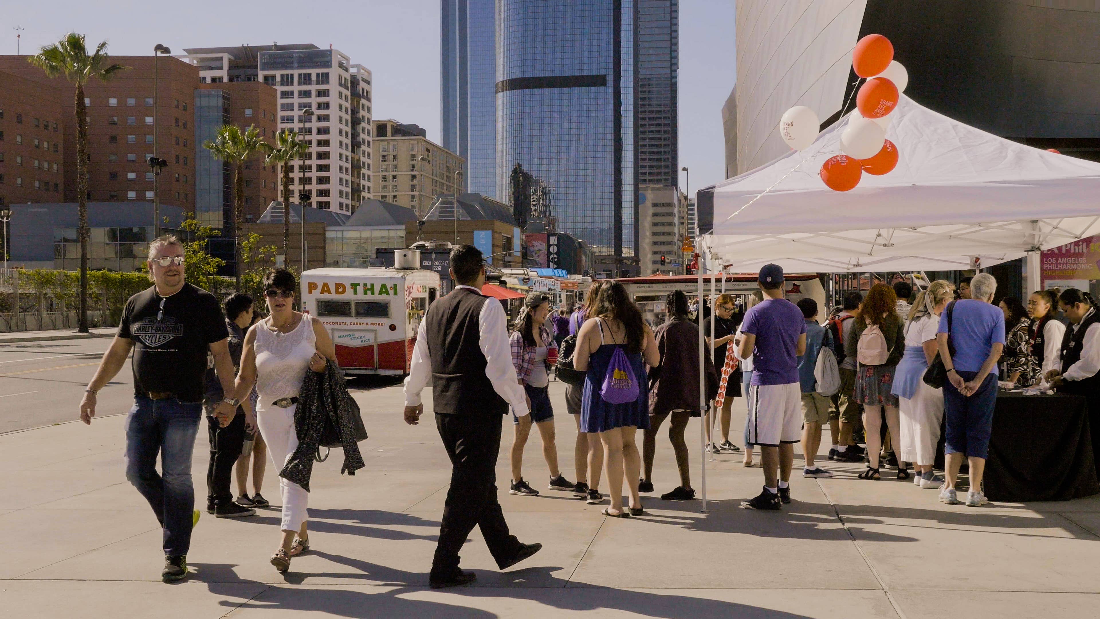 Crowd walks past food trucks and white event tents beneath tall downtown skyscrapers.