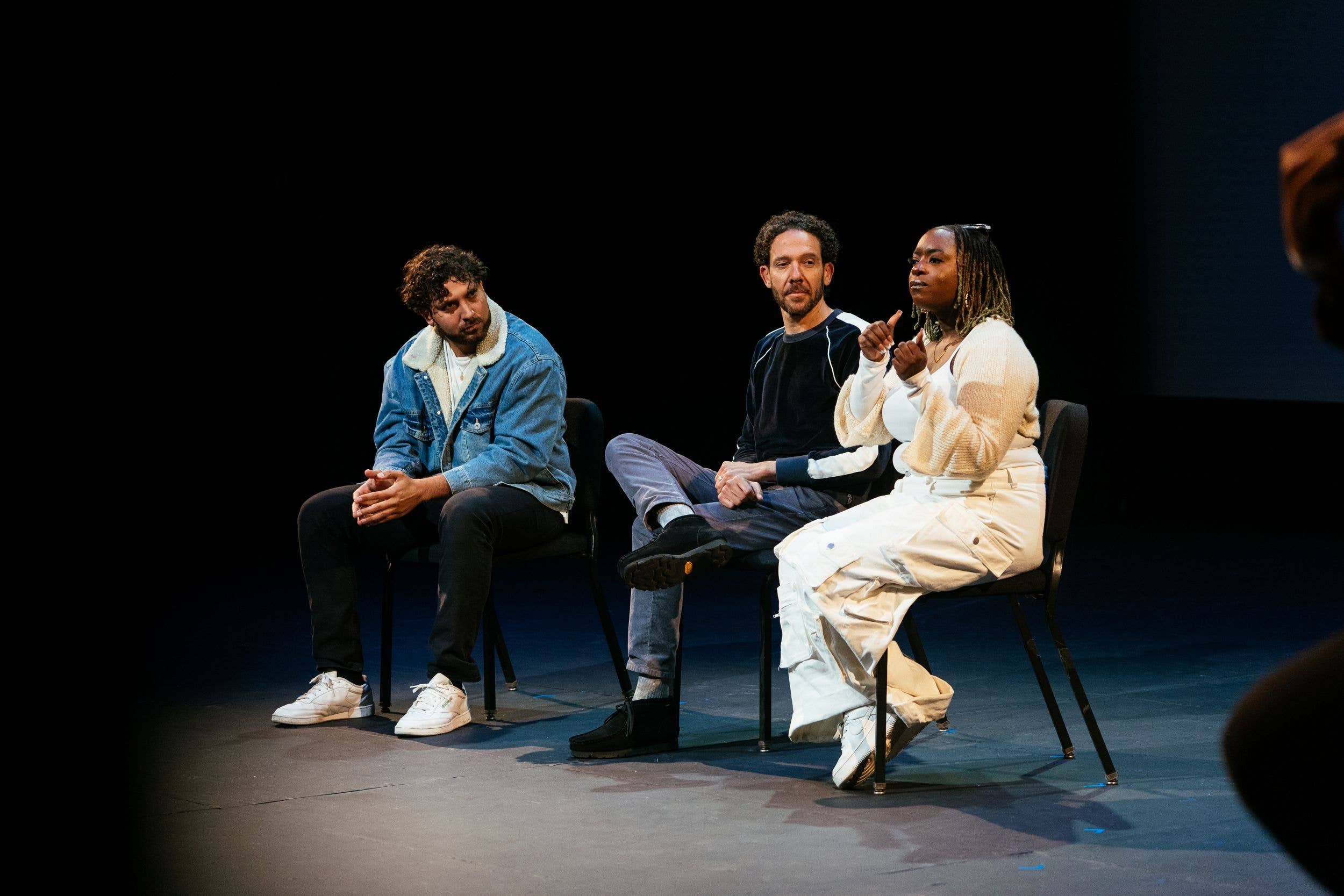 Three people seated on stage, one woman in white speaking and gesturing while two men listen.
