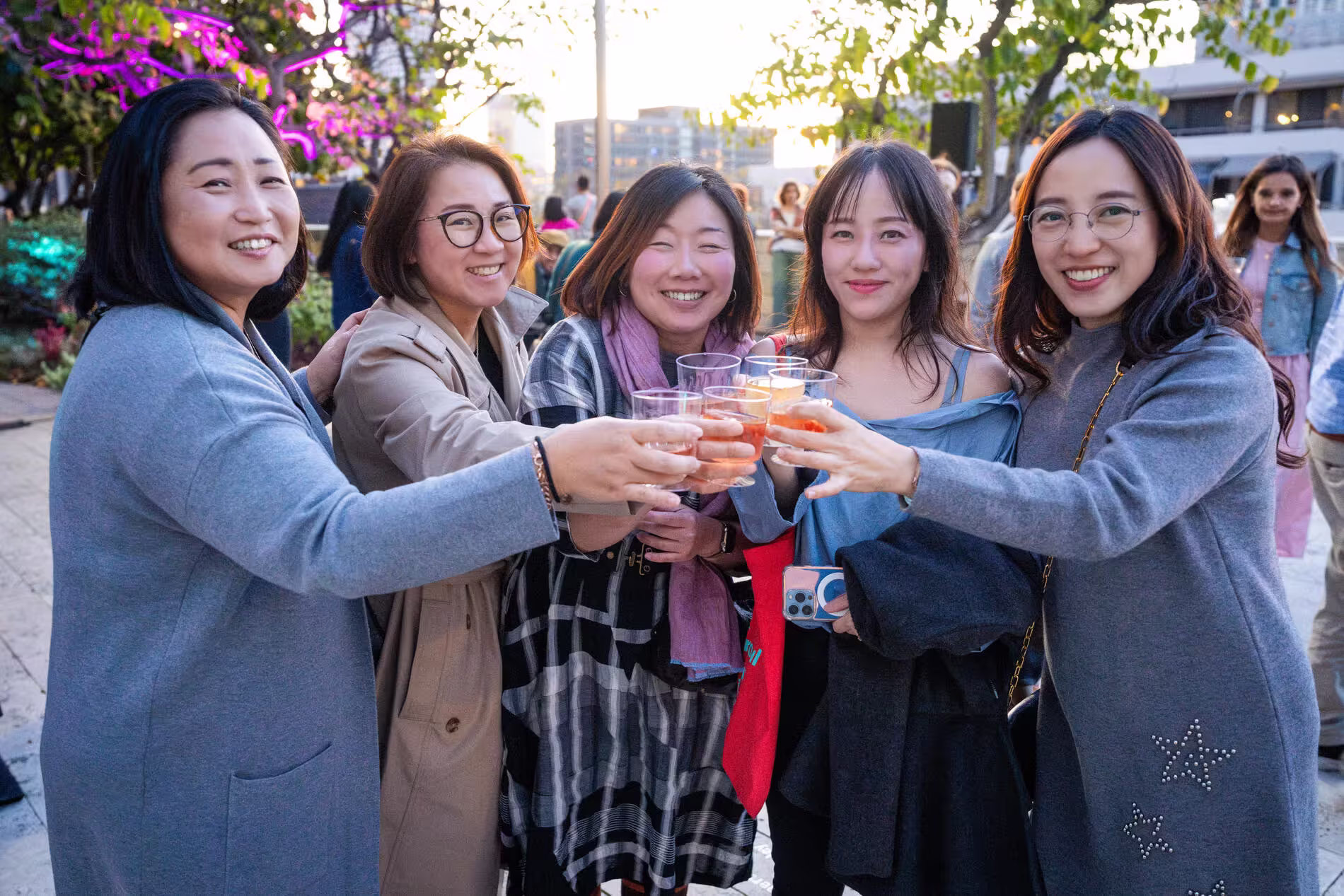 Group of five friends smiling and clinking glasses outdoors at sunset with colorful lights and city buildings behind