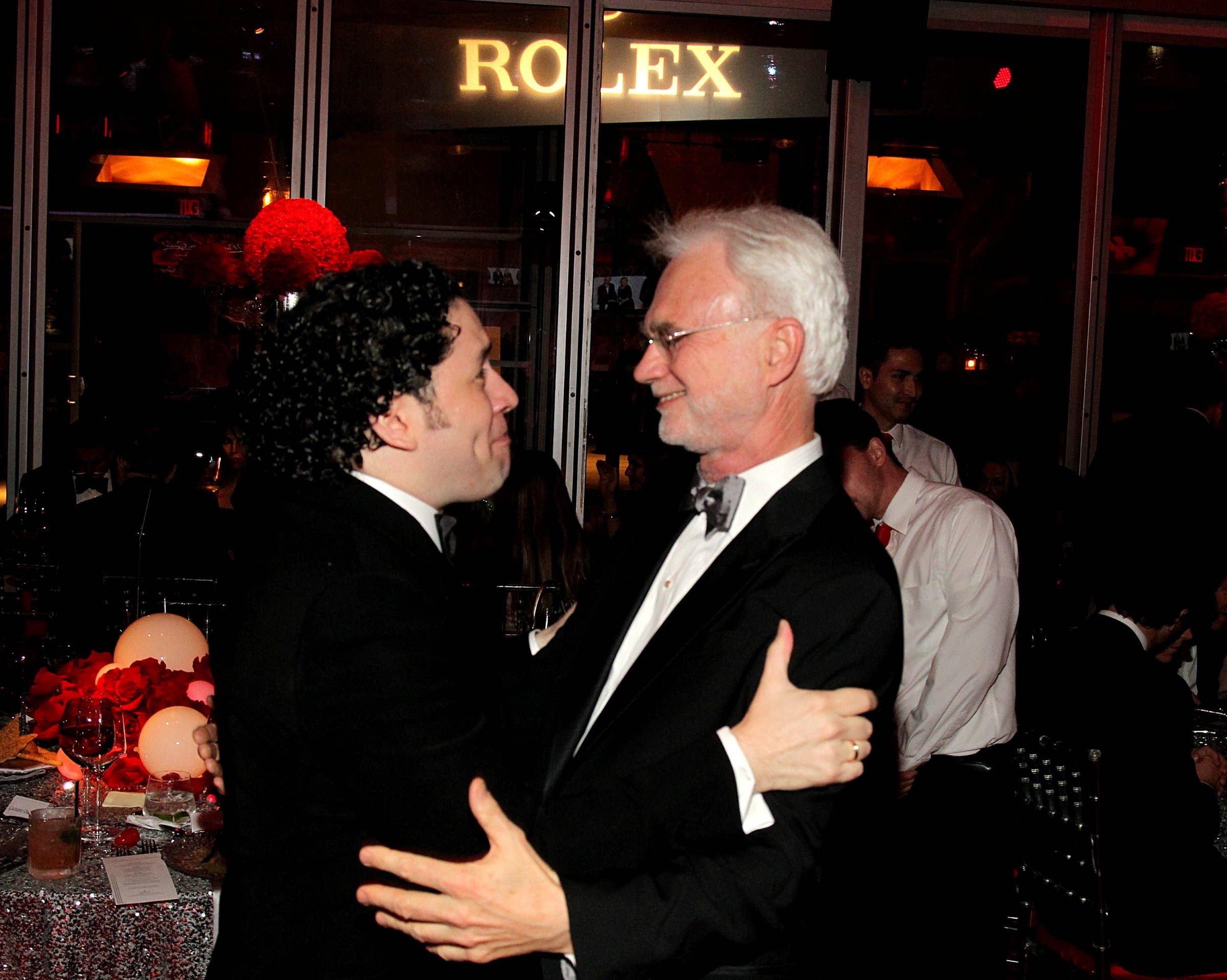 Two men in formalwear smile and embrace at a nightclub bar with a ROLEX sign in the background.