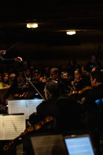 Conductor leads a packed orchestra, gesturing with a baton in a dim concert hall lit by stage lamps.