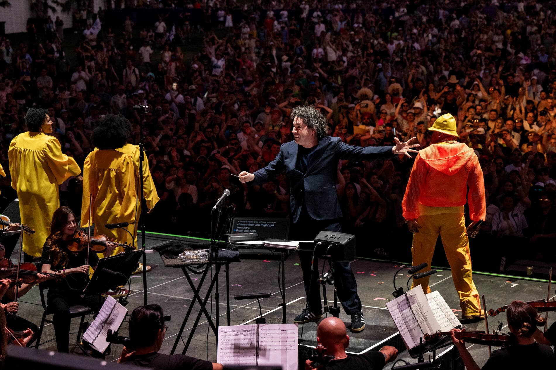 Musician in blue blazer welcomes crowd during concert; bandmates in yellow and orange stand at front of stage.