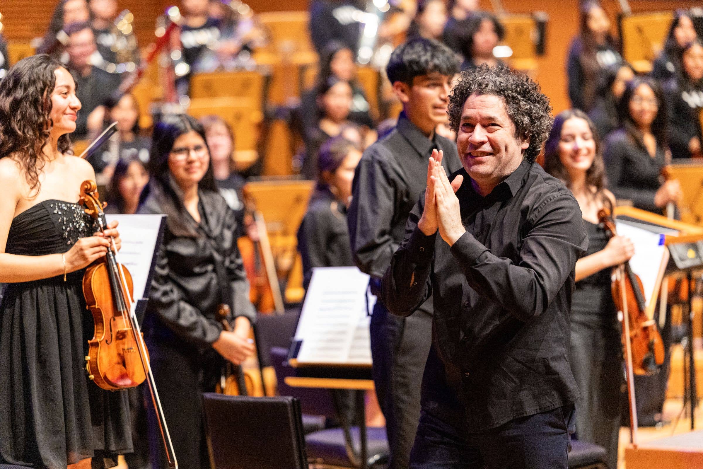 Curly-haired conductor smiles and applauds at a live orchestra performance in a concert hall, with musicians seated behind
