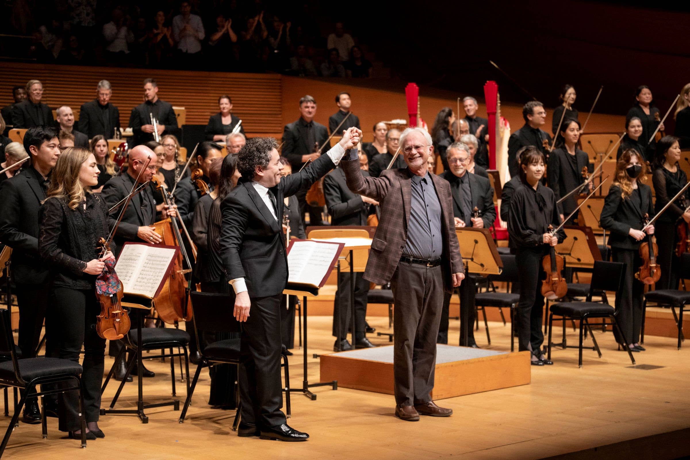 Conductor and orchestra musicians onstage, smiling and applauding after performance, with harps and string players.