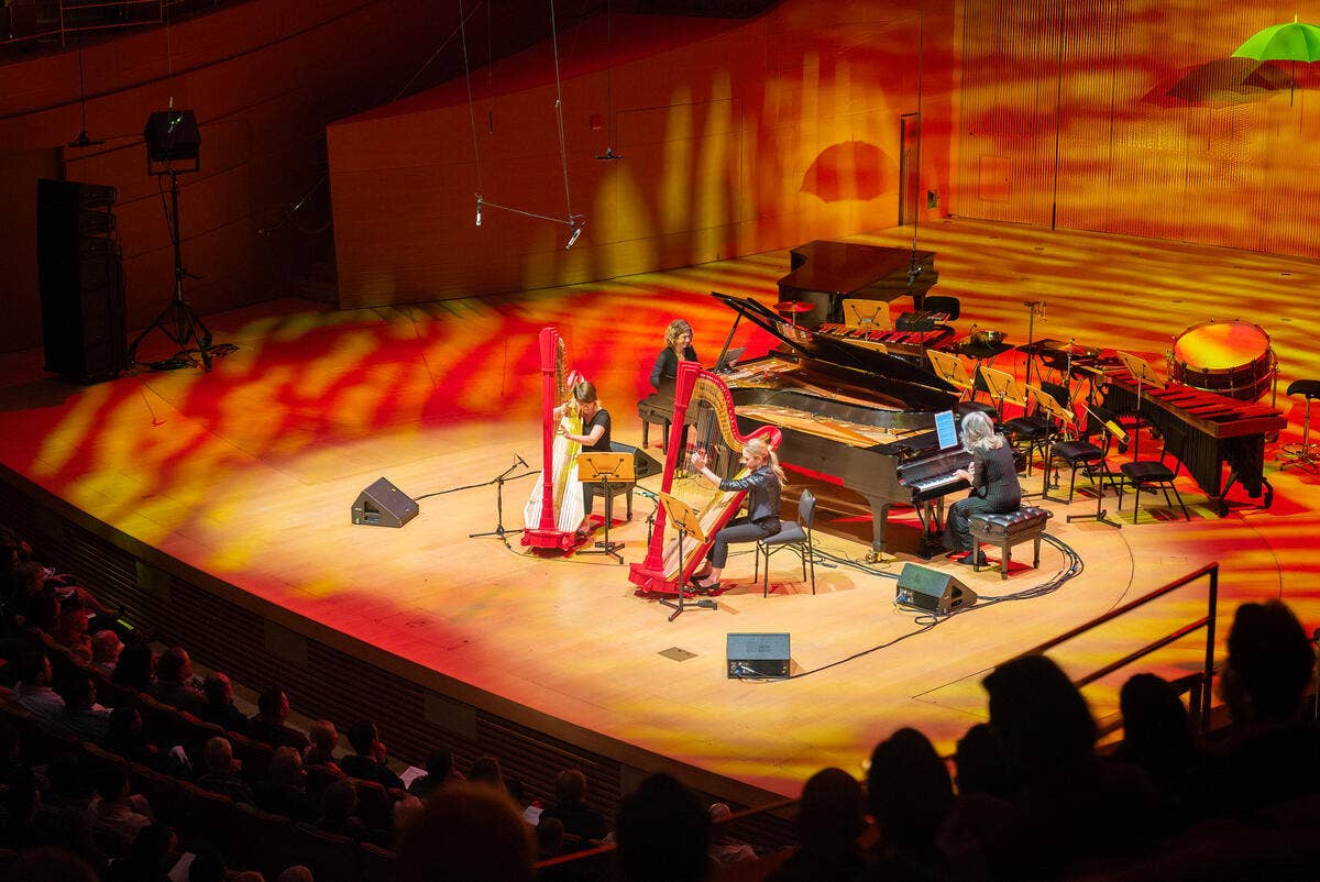 Musicians play piano and percussion on a theater stage with dramatic red and orange lighting, audience in foreground