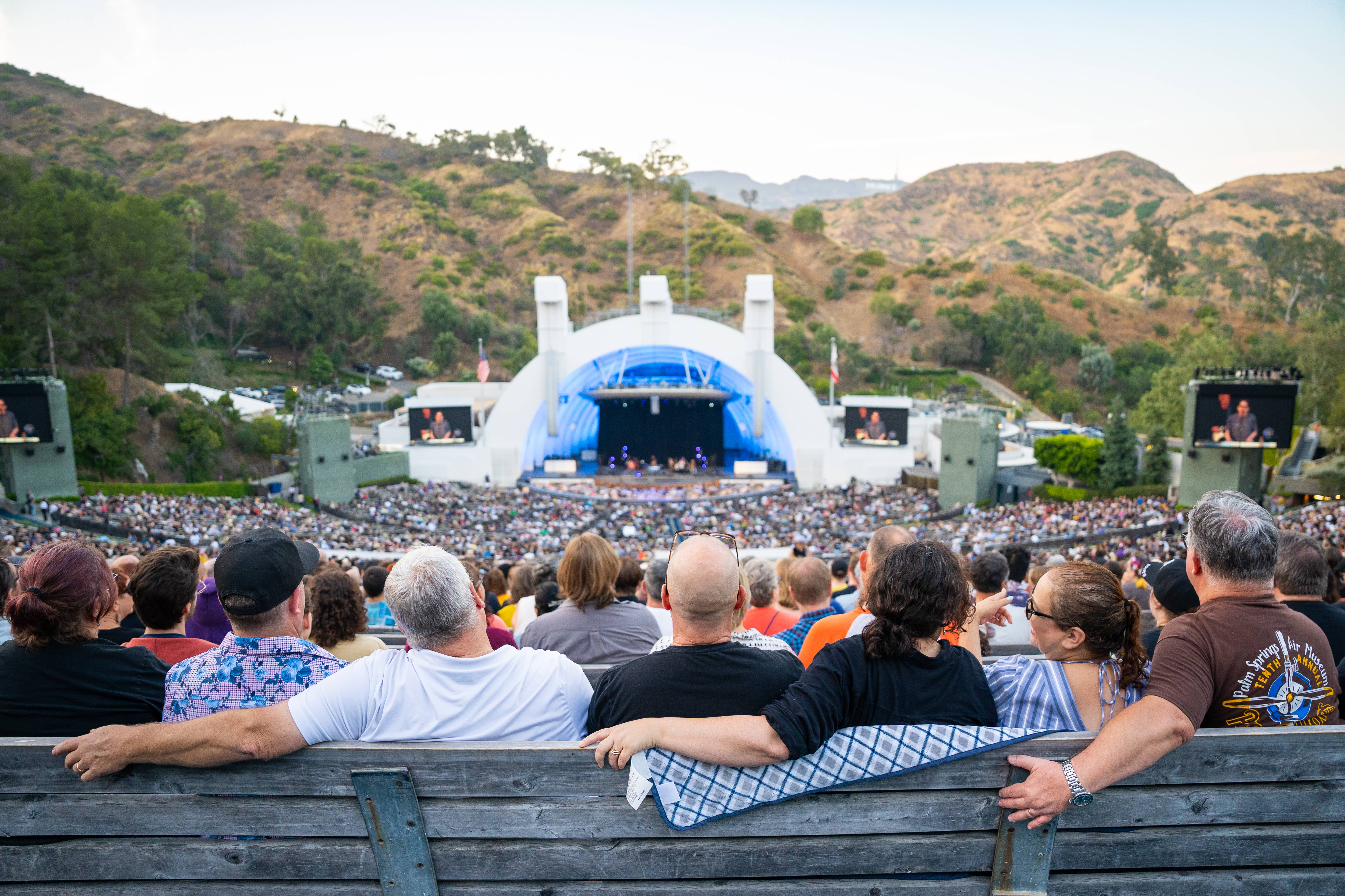 Crowd watching a live concert on an outdoor stage with blue lighting, surrounded by hills and trees at dusk