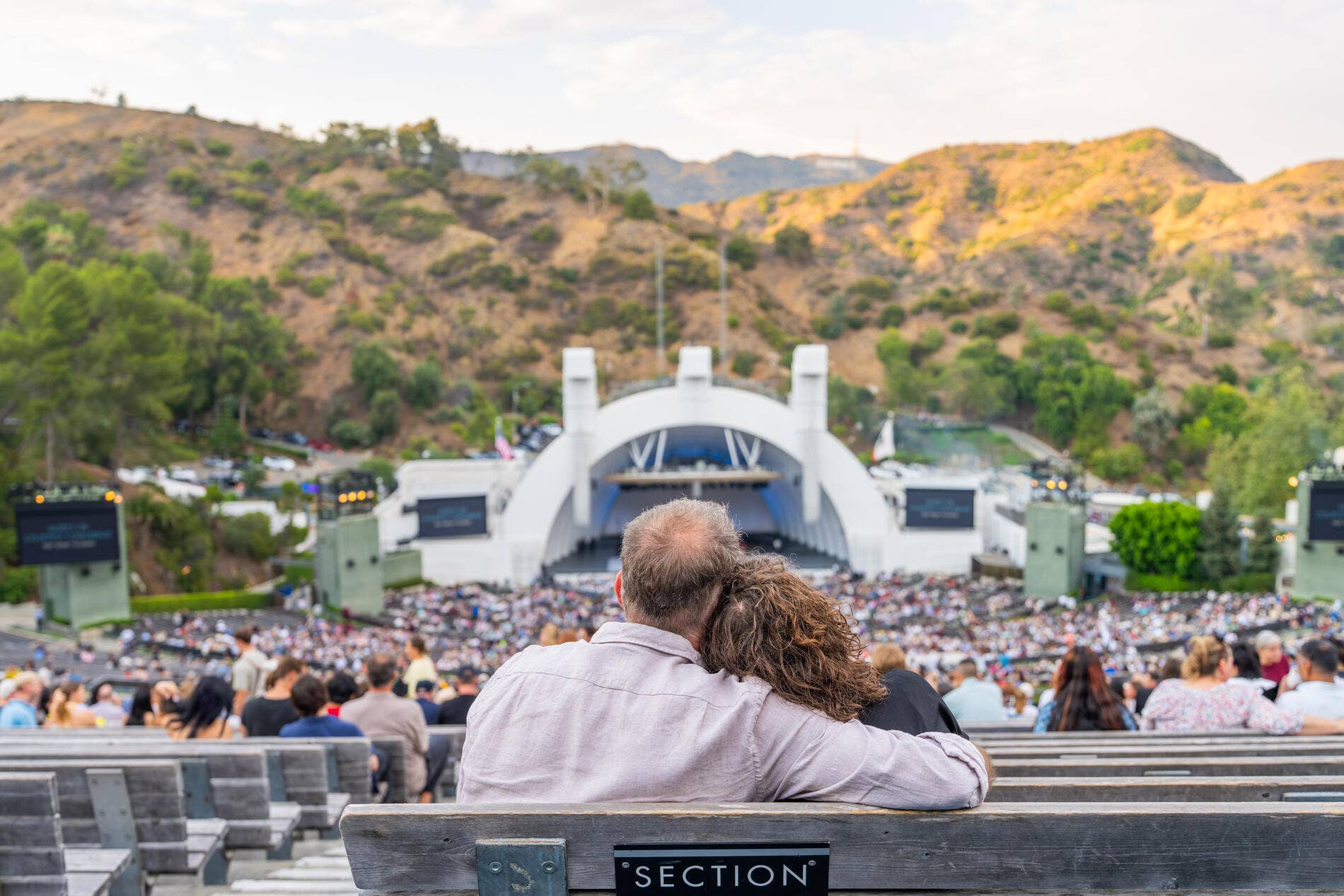 Couple watching a live concert from stadium seats, with a stage and sunlit hills in the background.