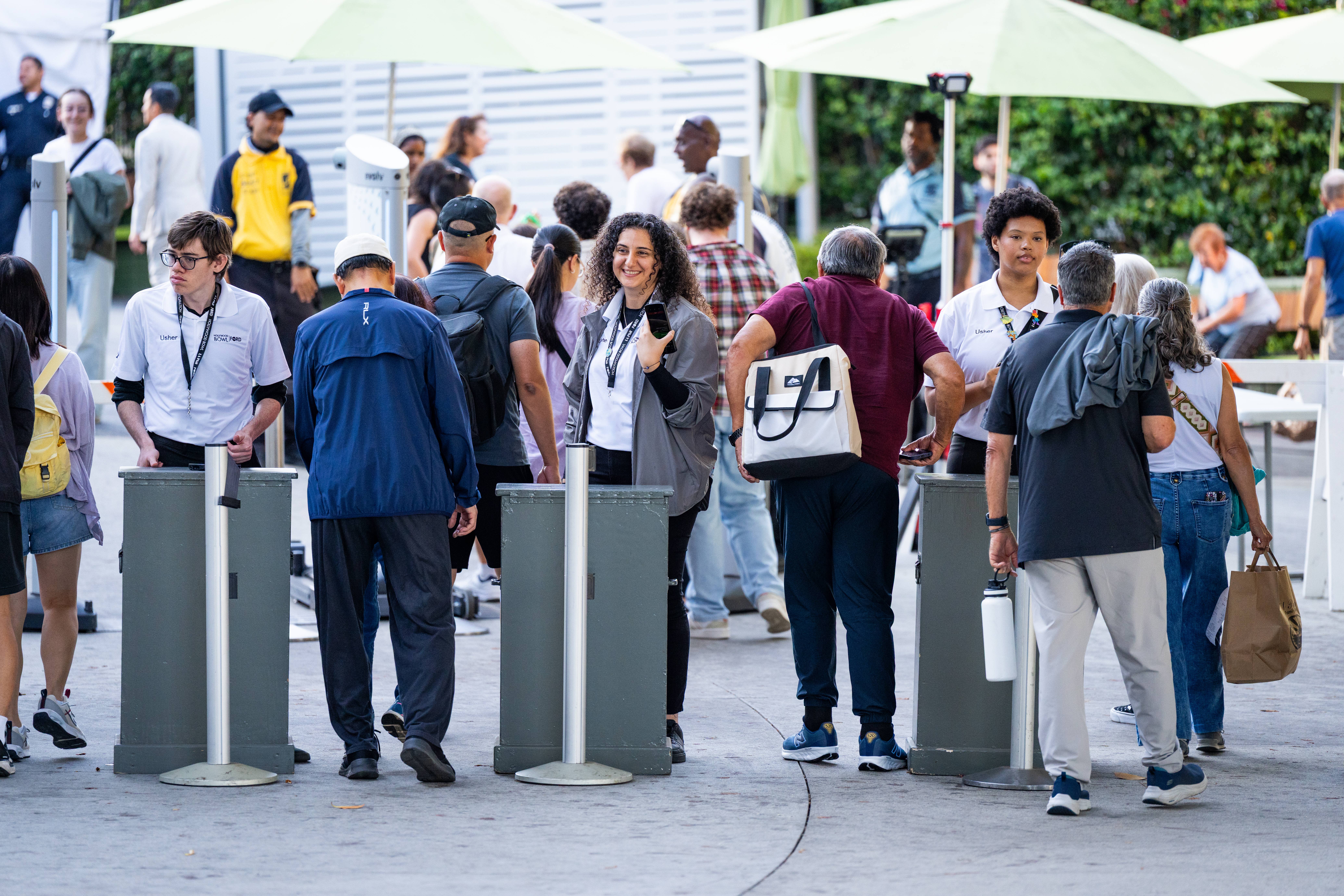 Crowd of attendees wearing lanyards walks between outdoor trash bins under white event umbrellas