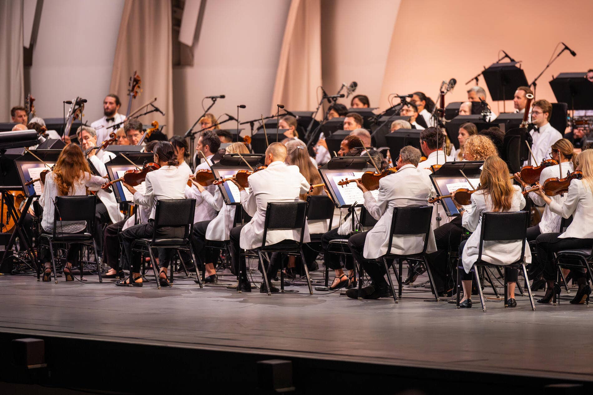 Orchestra musicians in white attire play violins and other instruments on a concert stage with microphones and speakers.