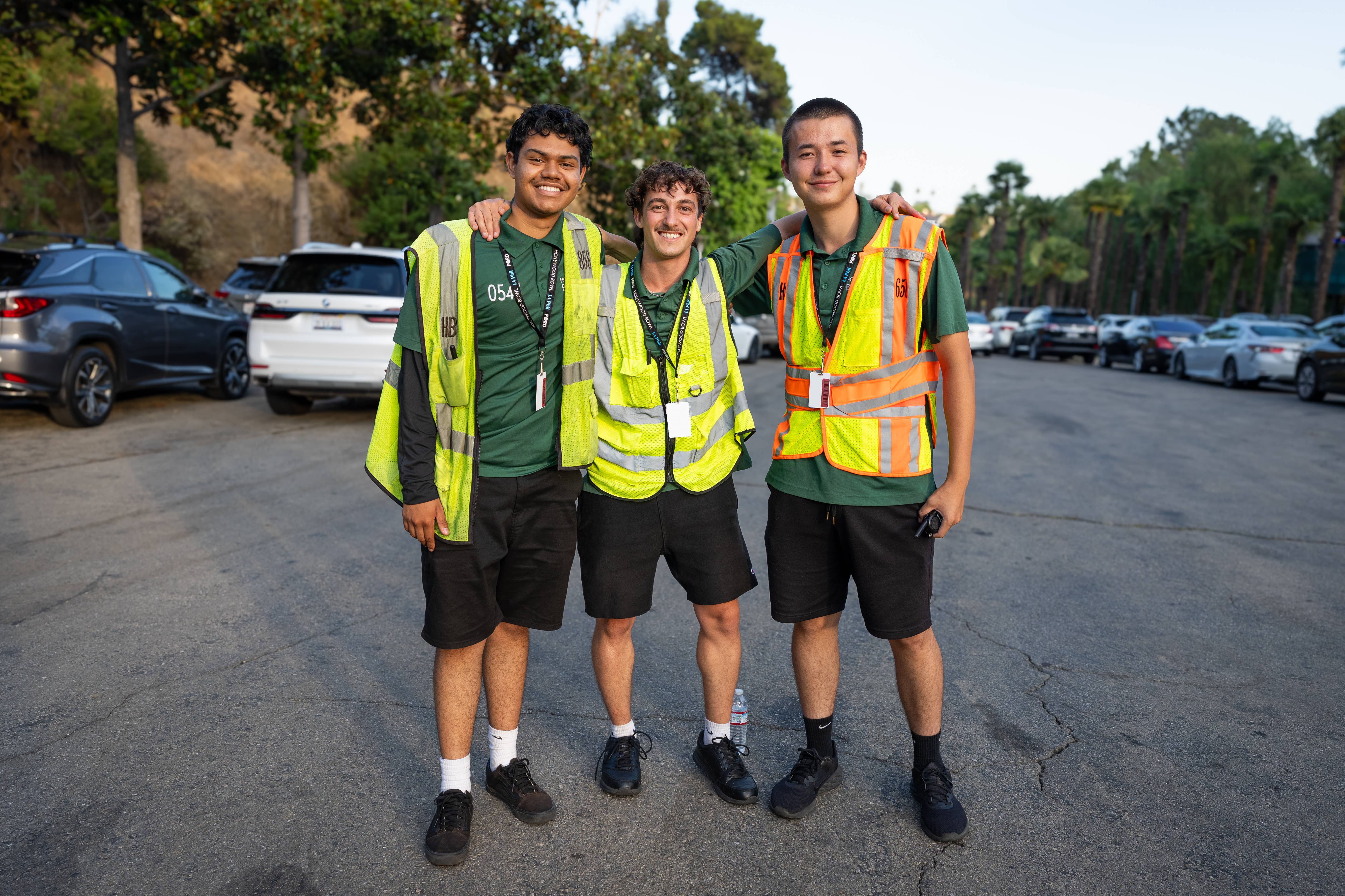 Three workers in high-visibility vests stand together outdoors in a parking lot, smiling at the camera.