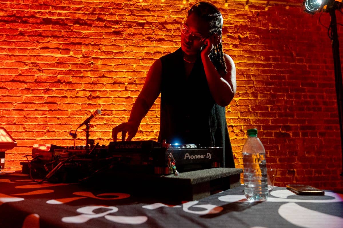 DJ standing at a Pioneer DJ controller, talking into a microphone with a red-lit brick wall behind him.