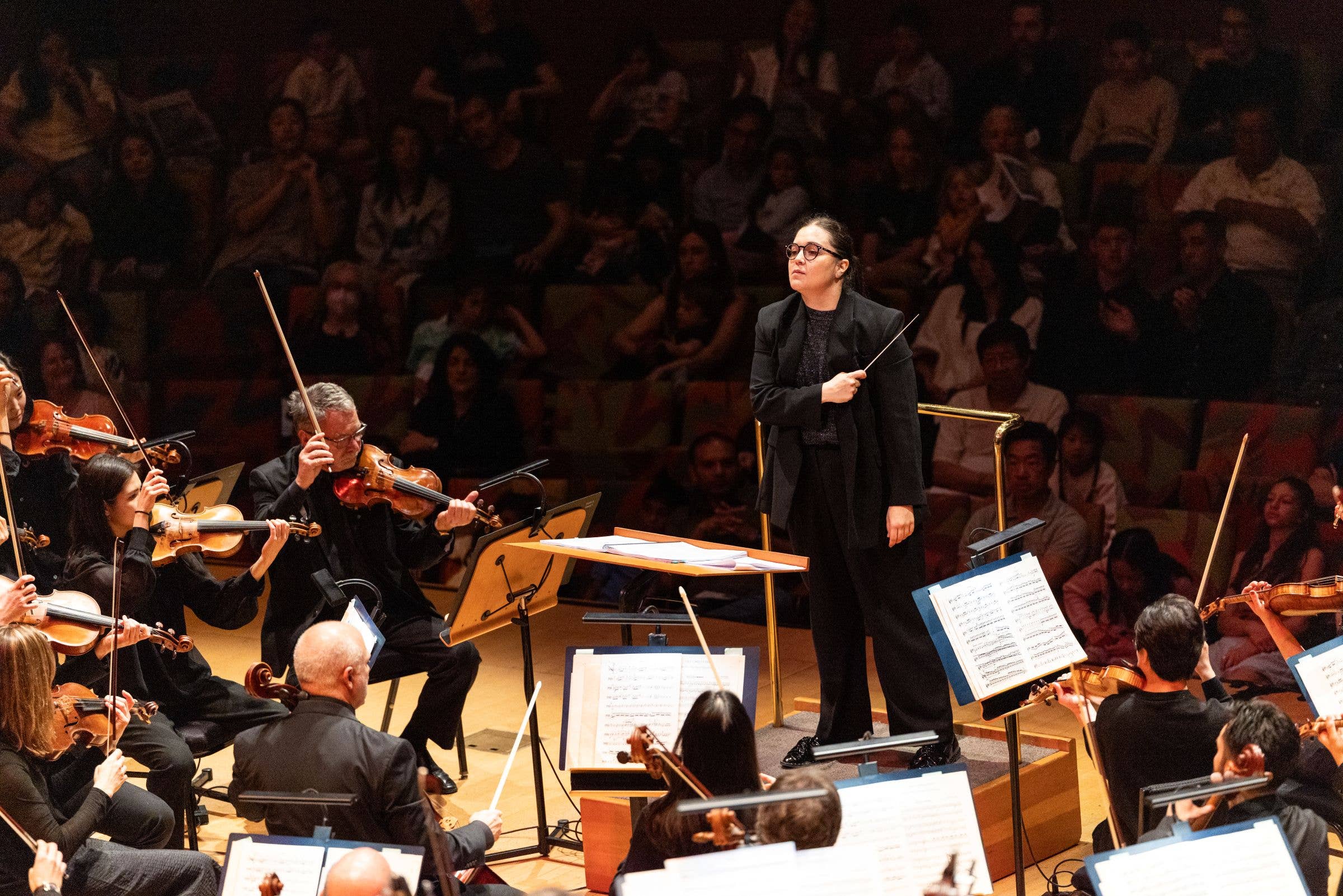 Female conductor with baton leading an orchestra, musicians seated with violins and sheet music in a concert hall