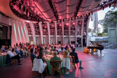 Guests seated at an outdoor gala dinner on a stage under red-lit lighting rig and piano performance area.